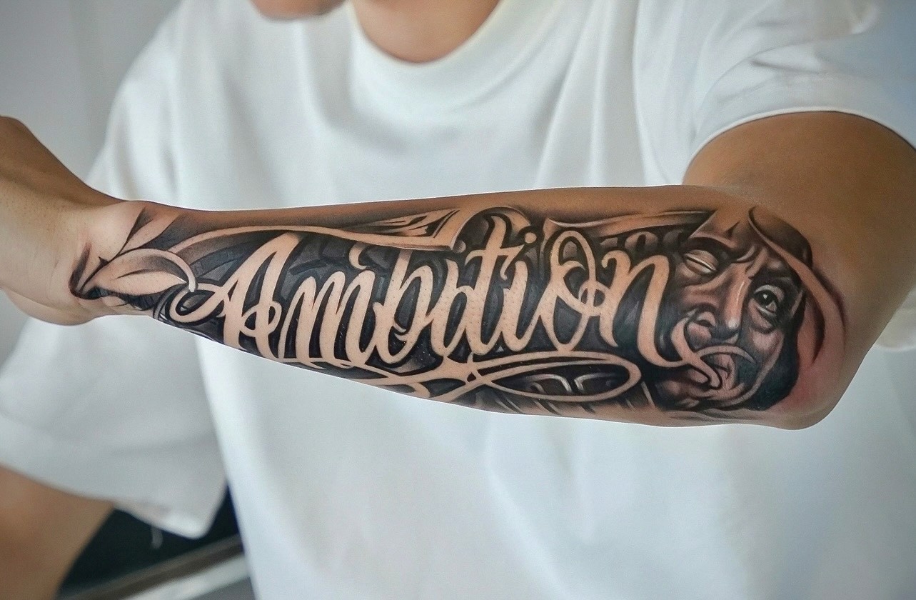 Chicano style script lettering tattoo reading Ambition on a forearm