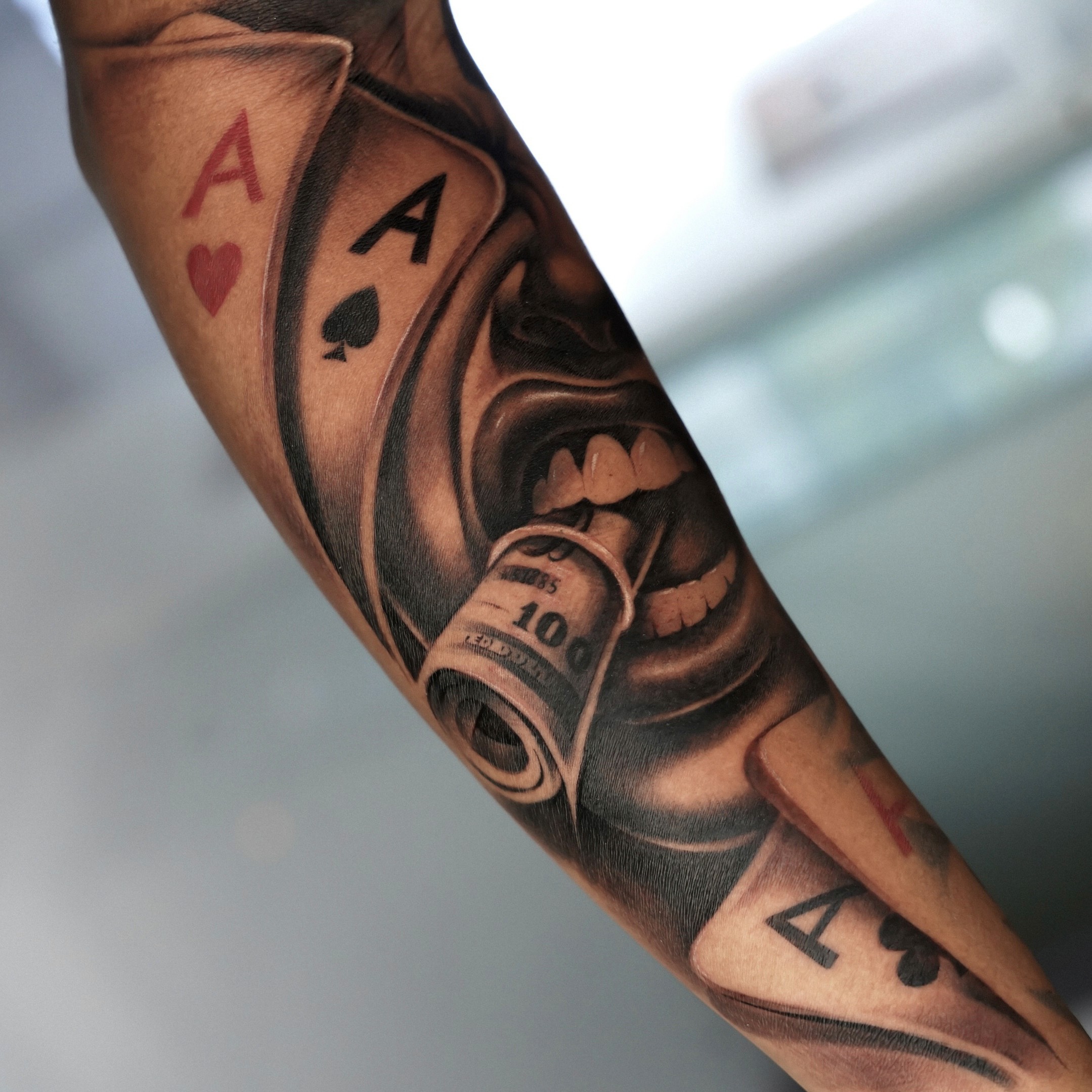 Forearm tattoo of a mouth biting a rolled hundred dollar bill flanked by ace cards